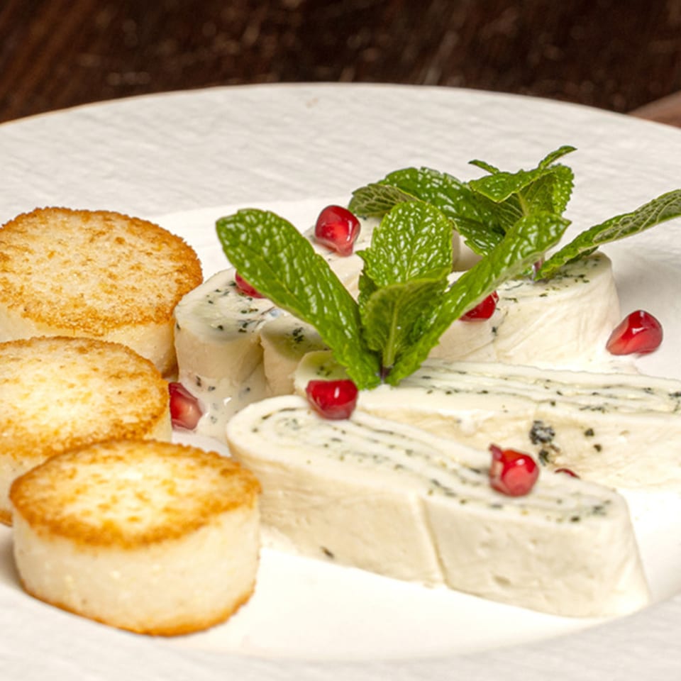 White cheese rolls with dark specks, garnished with mint and pomegranate seeds, beside three golden-brown patties.