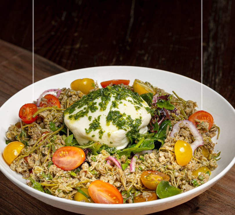 Burrata cheese with pesto, cherry tomatoes, red onion, and grains in a white bowl.